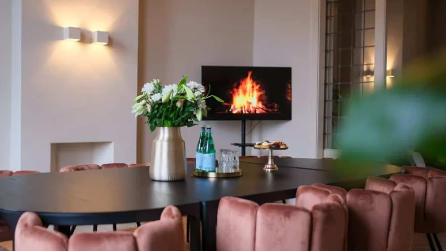 Een vergaderruimte met een lange tafel, roze stoelen en een haardvuur op het scherm