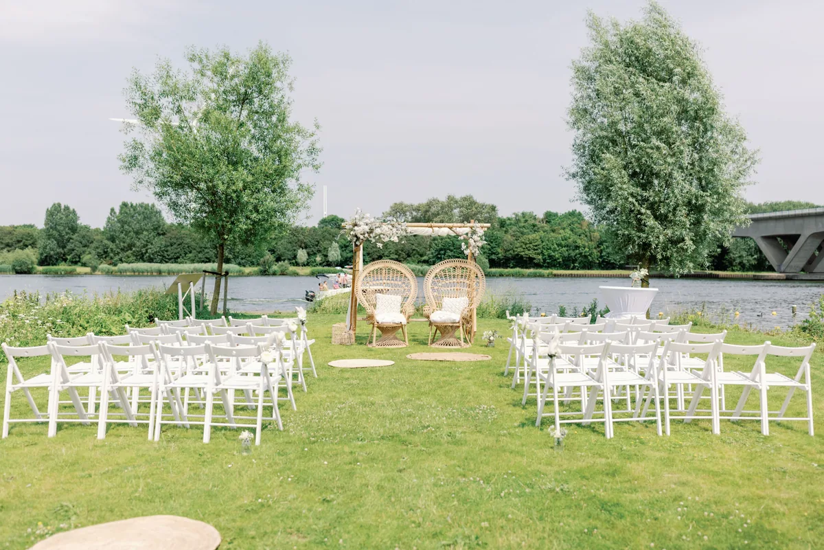 Witte stoelen en twee rotan stoelen onder een bloemenboog op het trouweiland aan het water