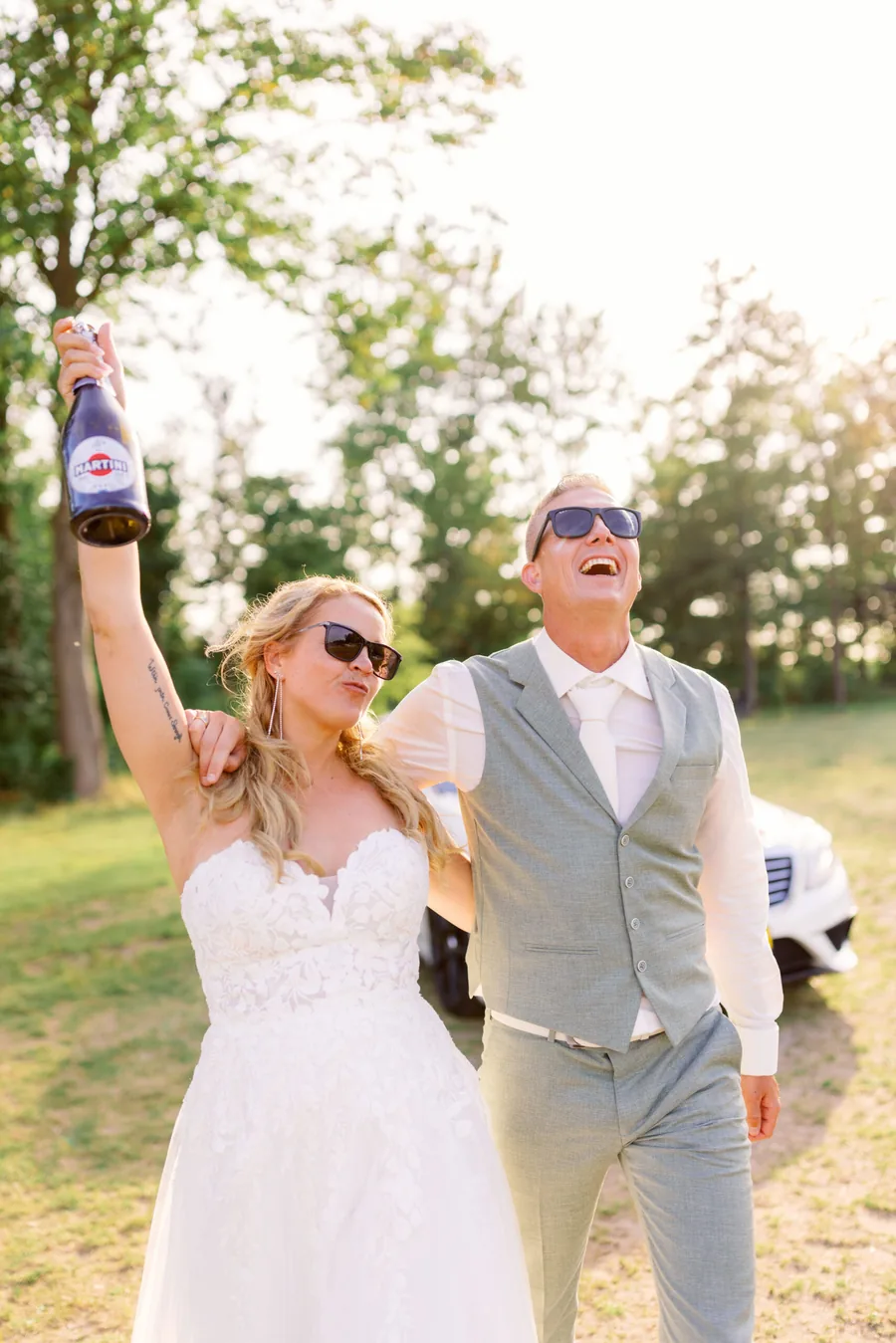 Een bruidspaar juicht met een fles in de hand op het gras aan het water