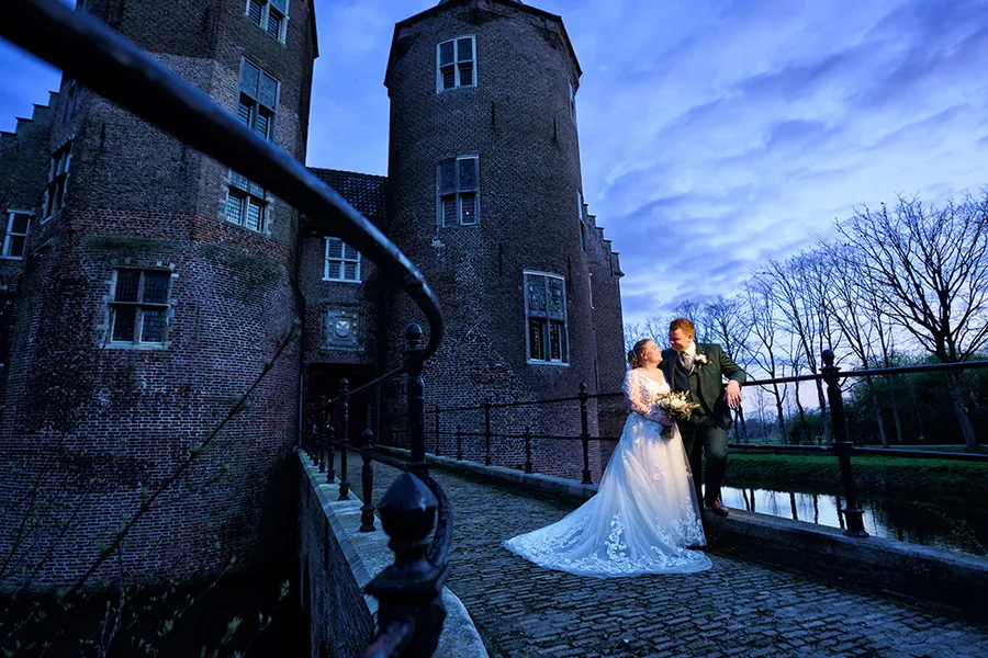 Een bruidspaar staat in het blauwe uur op de brug bij de kasteelmuur