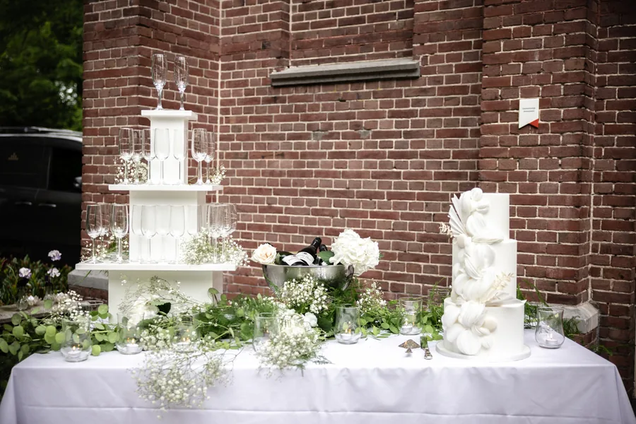 Een witte bruidstaart en een tweede taart op een gedekte tafel tegen een bakstenen muur